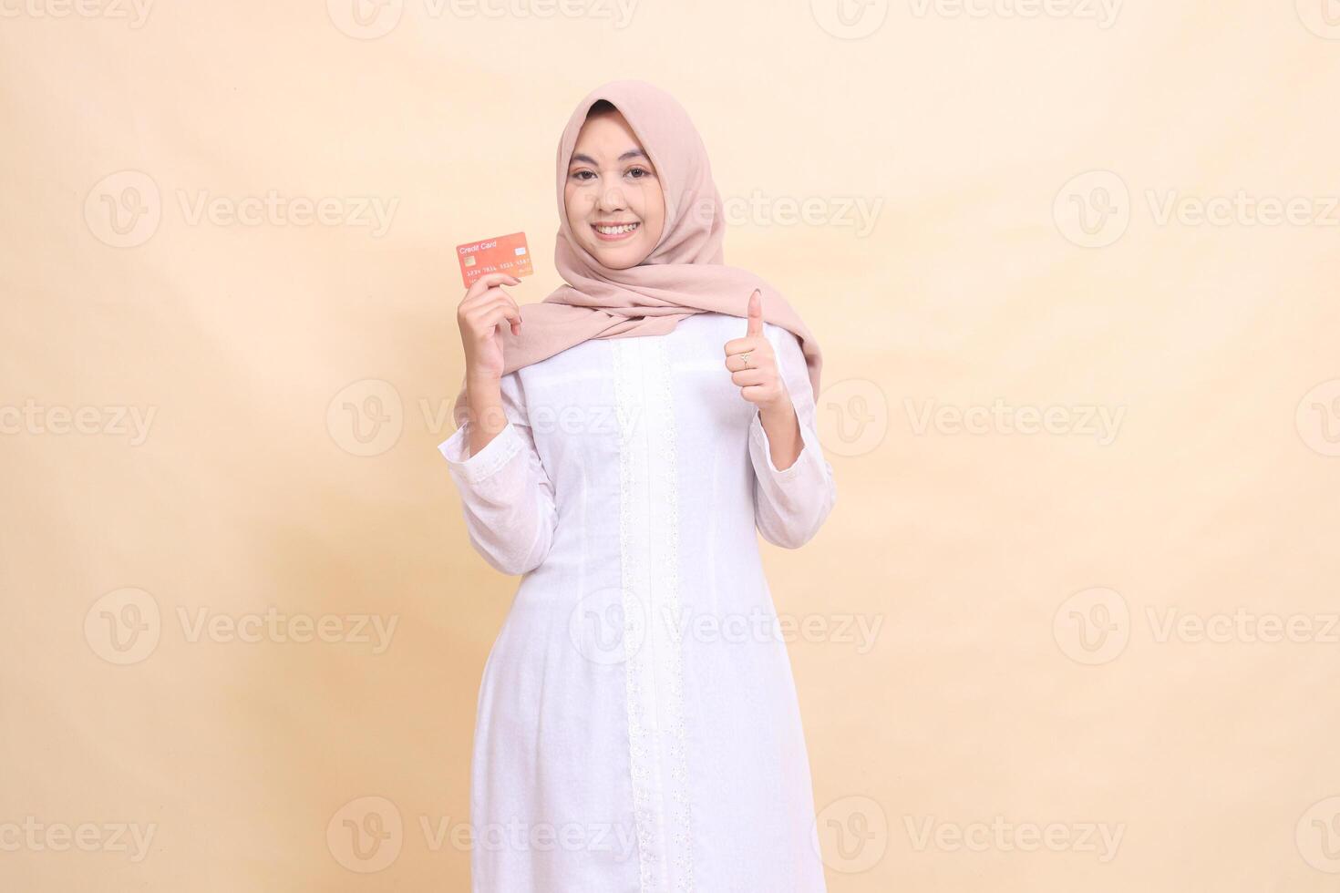 woman asian muslim wearing a hijab smiling cheerfully holding a debit credit card in her hand while giving a thumbs up gesture okay good good. Lifestyle, business and finance concept photo