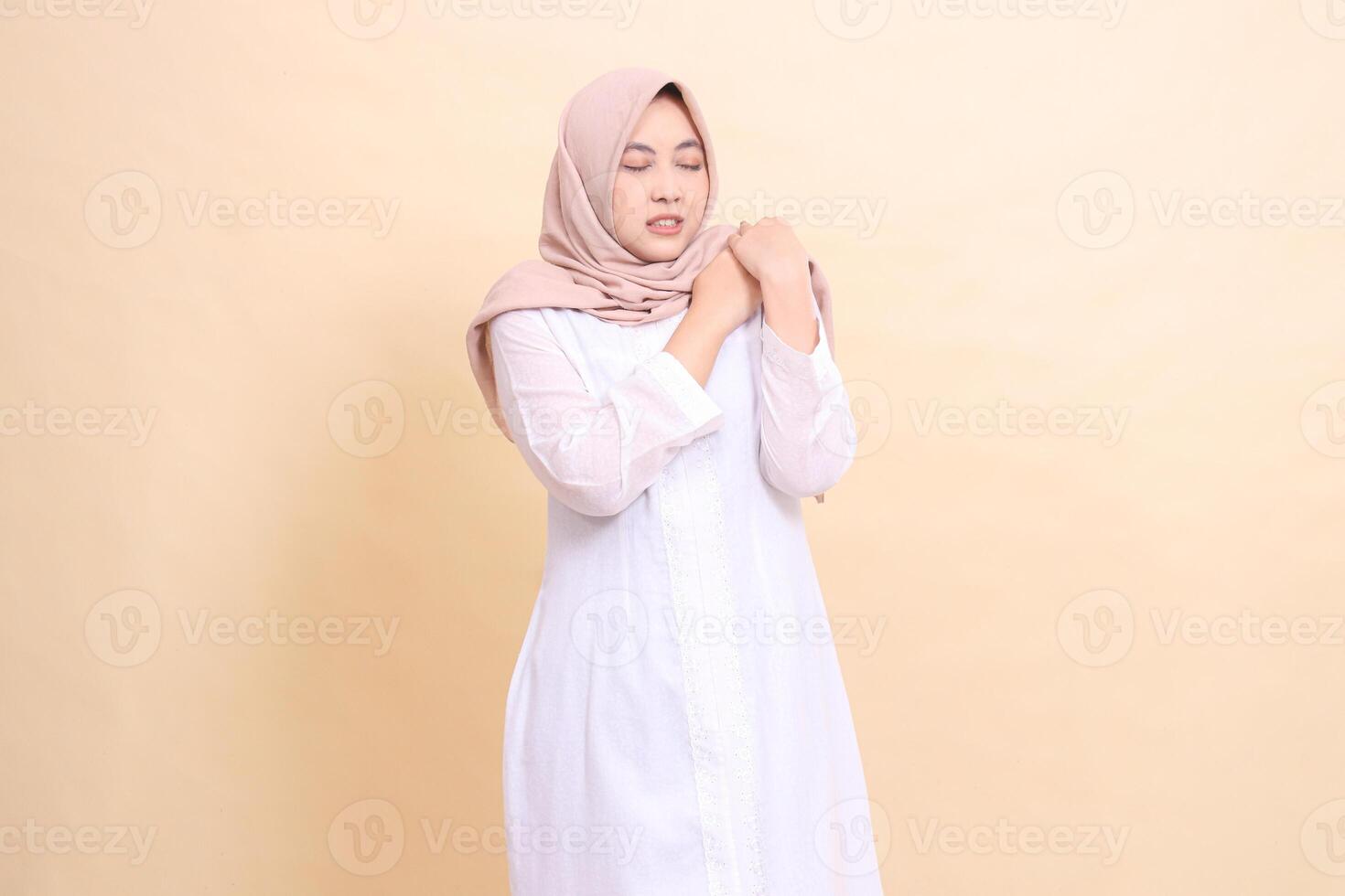 woman indonesia wearing a white hijab dress stands covering the word pain with both hands holding her shoulders shoulder pain. for health, care, education and science concepts photo