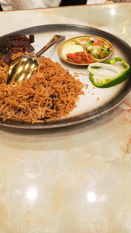 45 degree portrait of nasi kebuli kebuli rice in the middle with pickles, sauce and spoon, typical Arabic food made in Indonesia located on the table in closeup photo