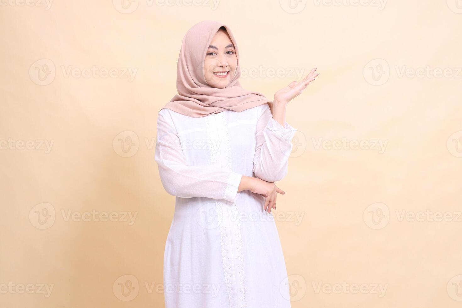 young Asian Muslim woman standing to the camera smiling with her arms crossed to the left. Fashion, pointer, business and promotion concept photo