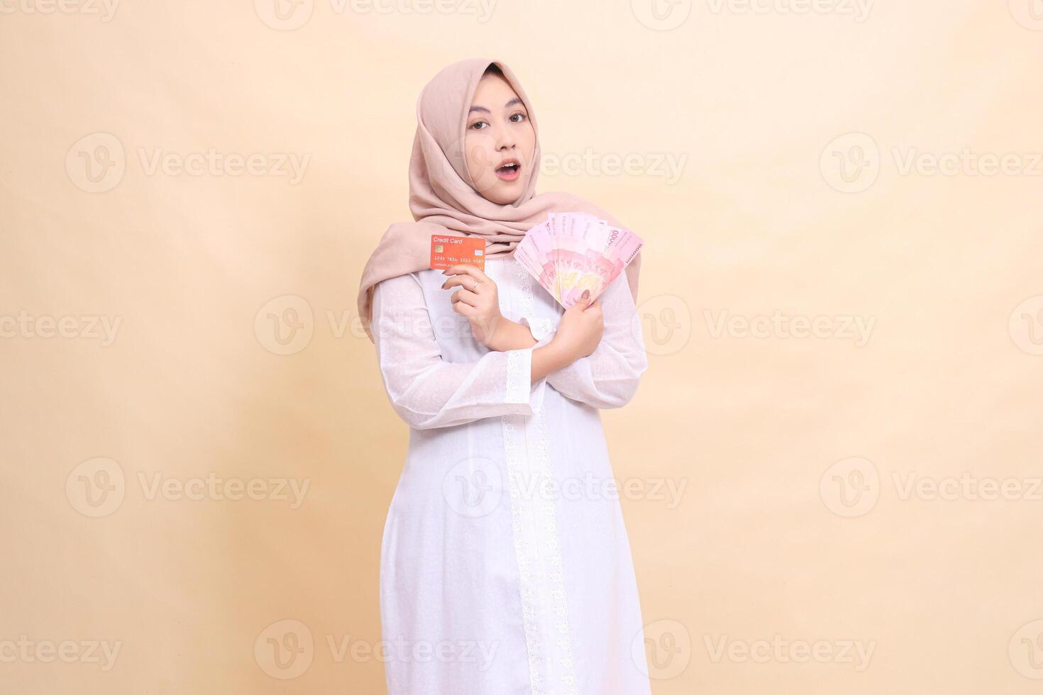 woman indonesia muslim in hijab shocked holding rupiah cash and credit card in crossed hands. Lifestyle, religious, business and finance concepts photo
