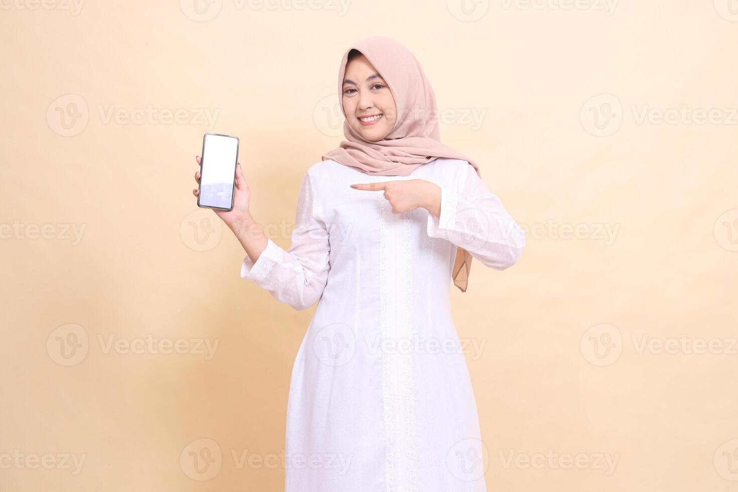 Young woman indonesia muslim wearing a hijab smiles cheerfully holding a smartphone gadget to the right in her hand while pointing at it. Lifestyle, business and commercial concept photo