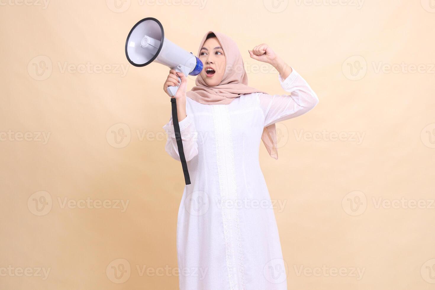 girl indonesia wearing hijab slanted to the right candidly shouts enthusiastically, holding a loudspeaker megaphone and clenching her fists upwards. Technology, broadcast and promotion concept photo