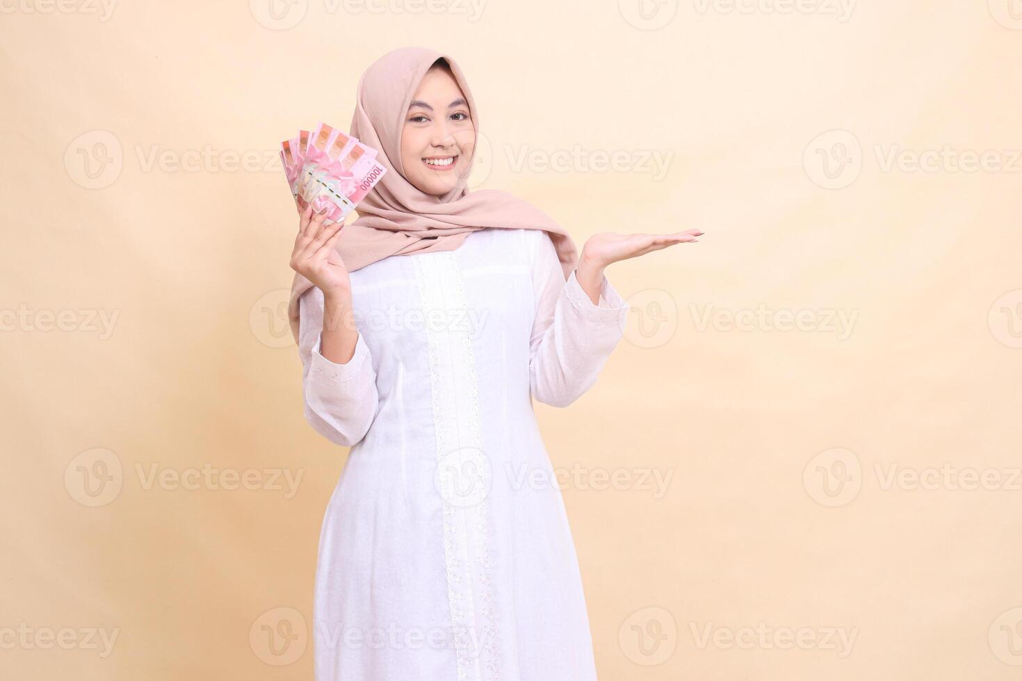 girl asia muslim in hijab smiles cheerfully holding rupiah cash in hand and presenting to the left. Lifestyle, religious, business and finance concepts photo