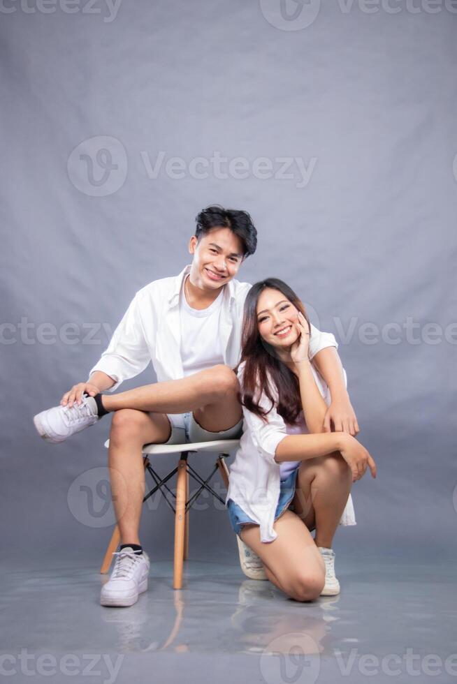 happy smiling man and woman posing sitting in front of the camera while their ladder hugs the woman indoors. fashion promotion couple shirt photo session in gray background studio