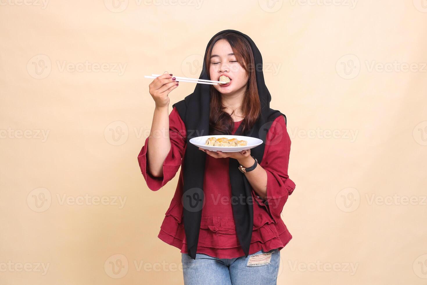 beautiful mature Asian woman wearing a red dress wearing a hijab smiling closing her eyes biting into dimsum using chopsticks and carrying a plate containing dimsum Chinese food photo
