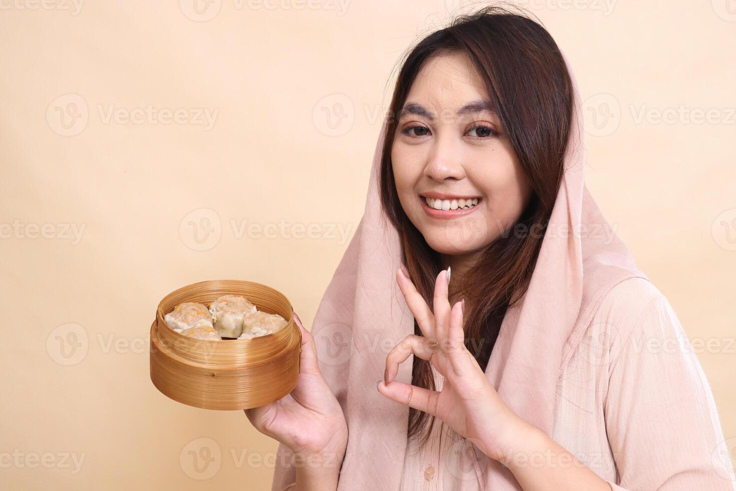 indonesia woman wearing a hijab tilted right to the camera smiling cheerfully with an okay good gesture and a wooden bowl to the side containing chopsticks and delicious and delicious dimsum photo