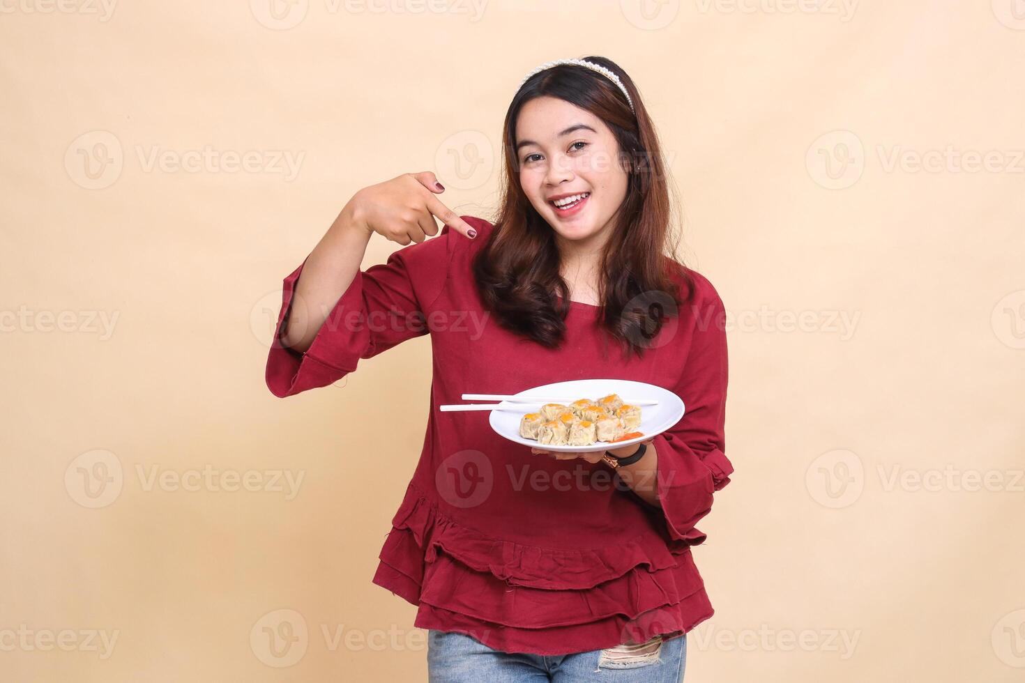 Beautiful young Asian woman in red dress cheerfully carrying a plate containing dimsum Chinese food, chopsticks and sauce while pointing at it. used for food, health and lifestyle content photo