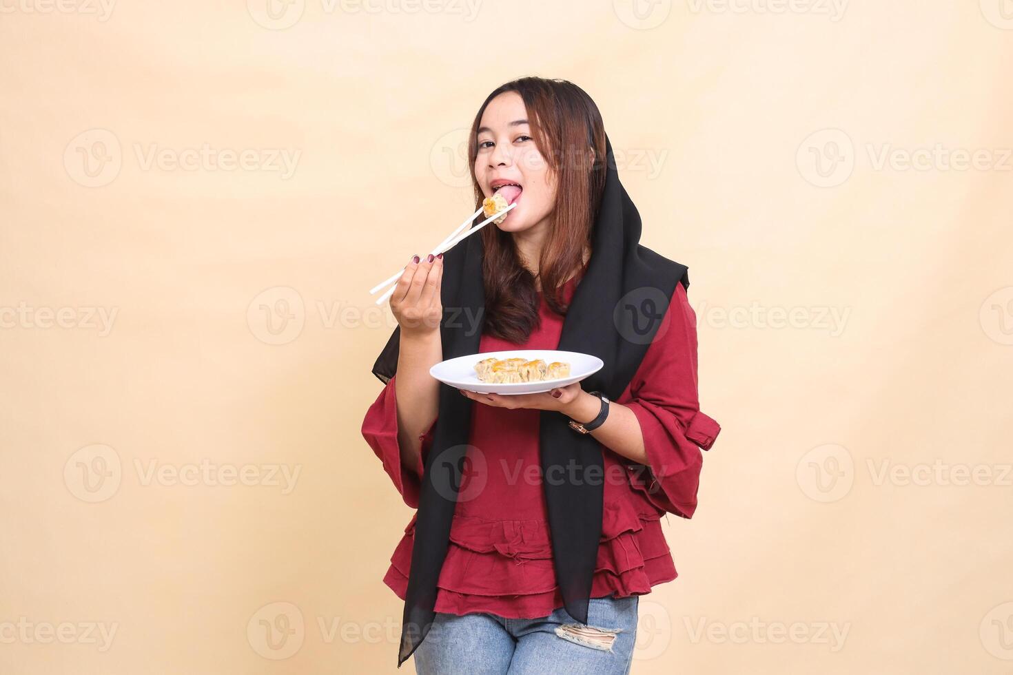 Beautiful mature indonesia woman in red dress wearing a hijab licks dimsum with chopsticks and brings a plate containing dimsum Chinese food to the front. used for food, health and culinary content photo