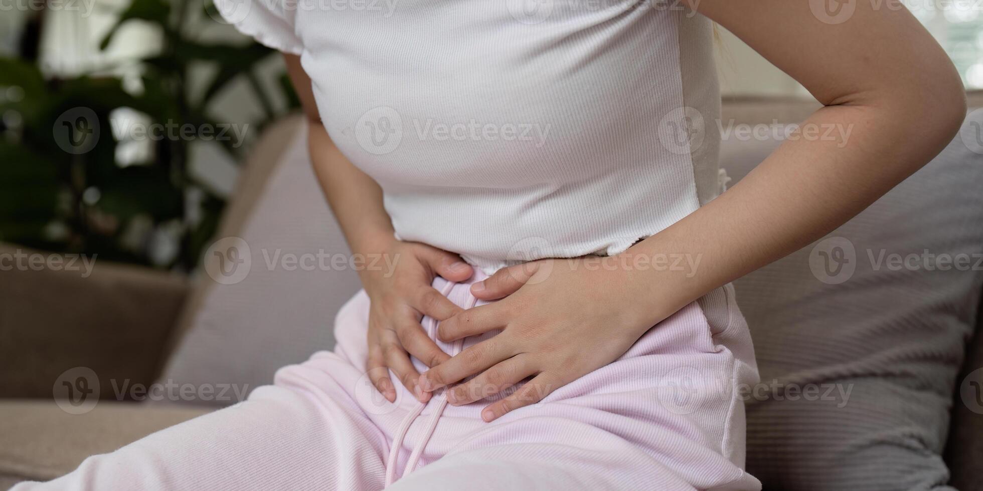 Woman Experiencing Stomach Pain While Sitting on a Couch in a Home Setting, Clutching Her Abdomen in Discomfort photo