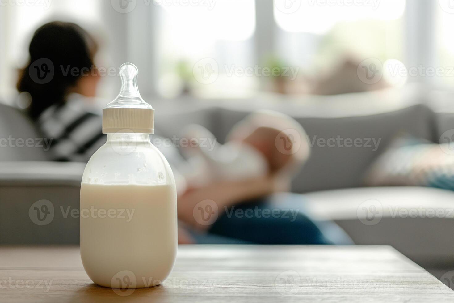 Baby feeding bottle with milk on a table in a bright room, in the background, out of focus, a mother holds a newborn in her arms while sitting on the sofa photo