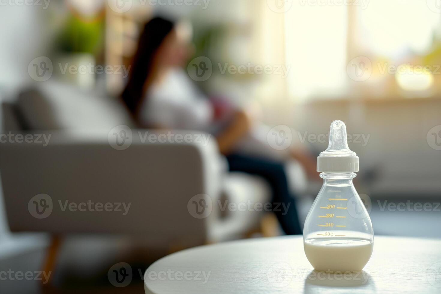 Baby feeding bottle with milk on a table in a bright room, in the background, out of focus, a mother holds a newborn in her arms while sitting on the sofa photo