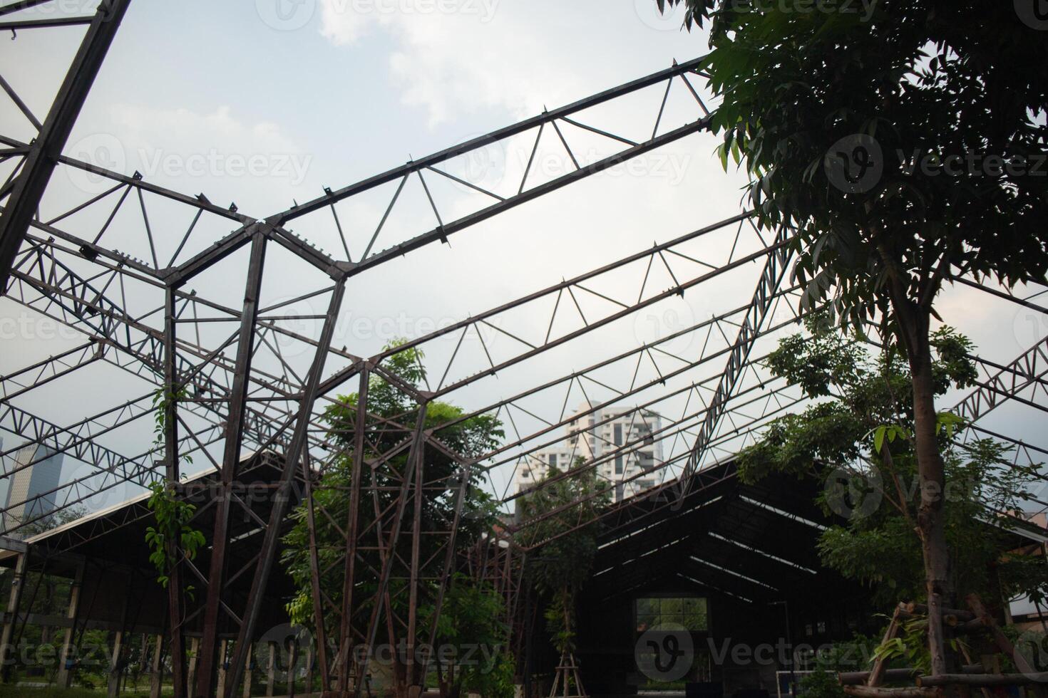 Abandoned greenhouse warehouse metal structure frame roof cover with tree and plant overgrown against blue sky in spring summer daytime photo