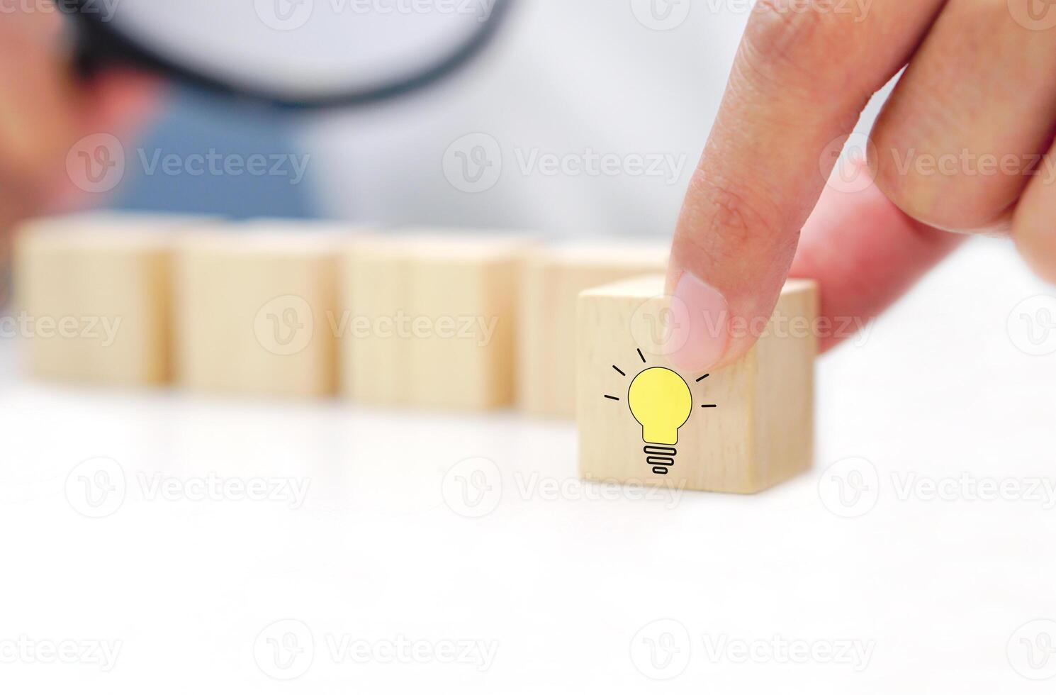 Hands holding a wooden cube with a light bulb icon symbol and blurred wooden blocks behind on a white background. Target of business, innovative idea and creative goals concept. photo