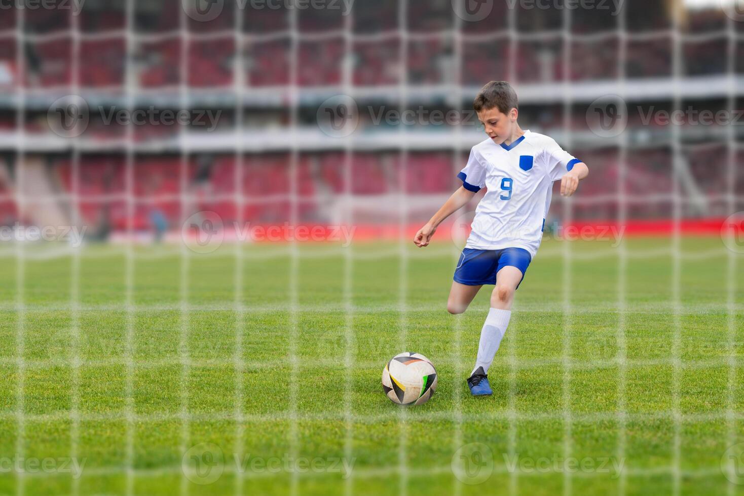 Young soccer player in a white dress shooting towards the goal on a modern stadium. View behind net photo