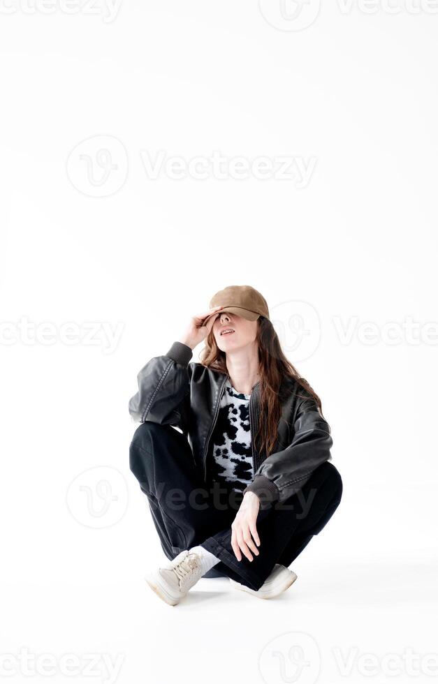 Young woman in a bomber jacket, jeans and a cap with braces poses while sitting in the studio, on a white background photo