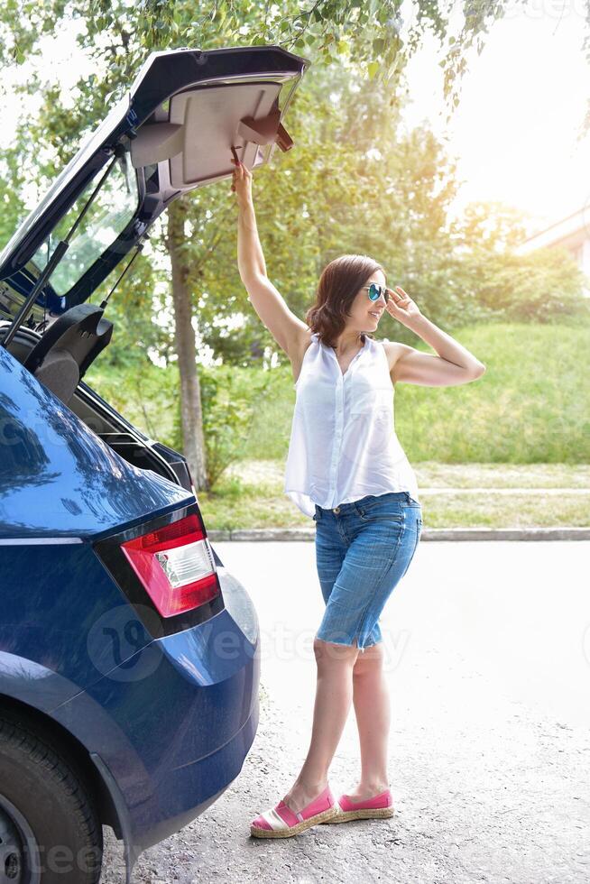 Smiling Caucasian woman in a white blouse and denim shorts putting her stuff bags into the car trunk. Sunny day photo