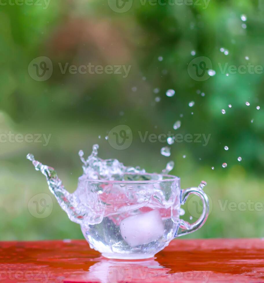 Splash of cool fresh water with ice cube in transparent glass cup in the table outoors in summer day. photo