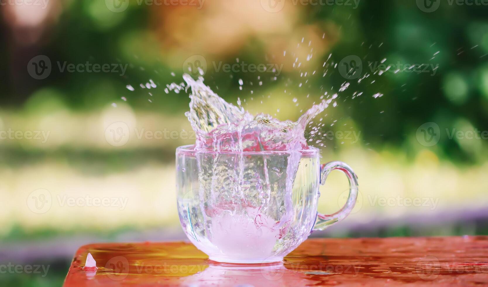 Splash of cool fresh water with ice cube in transparent glass cup in the table outoors in summer day. photo