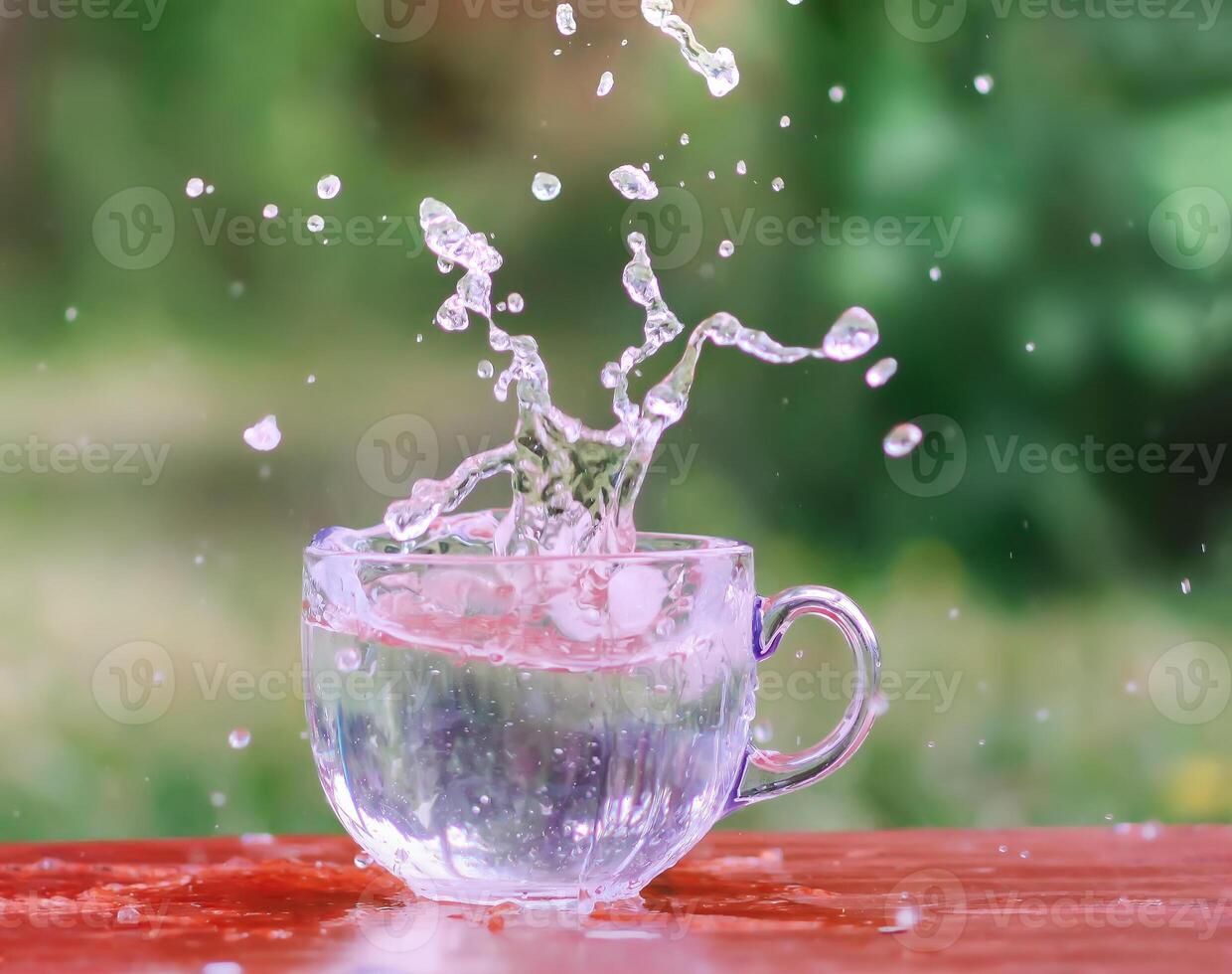 Splash of cool fresh water with ice cube in transparent glass cup in the table outoors in summer day. photo