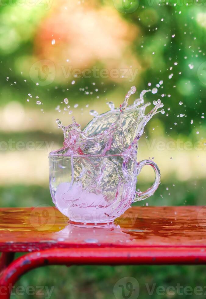 Splash of cool fresh water with ice cube in transparent glass cup in the table outoors in summer day. photo
