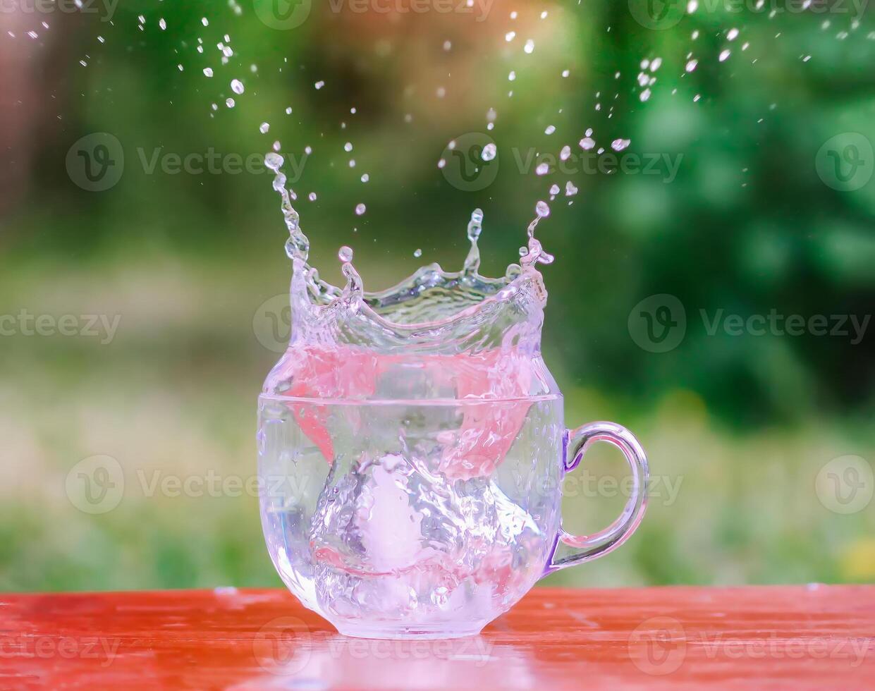 Splash of cool fresh water with ice cube in transparent glass cup in the table outoors in summer day. photo