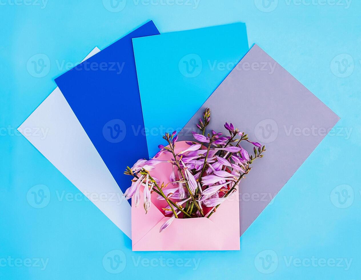 Pink paper envelope with fresh bright garden flowers and empty paper sheets on light blue background. Festive floral template. Greeting card design. Top view. photo