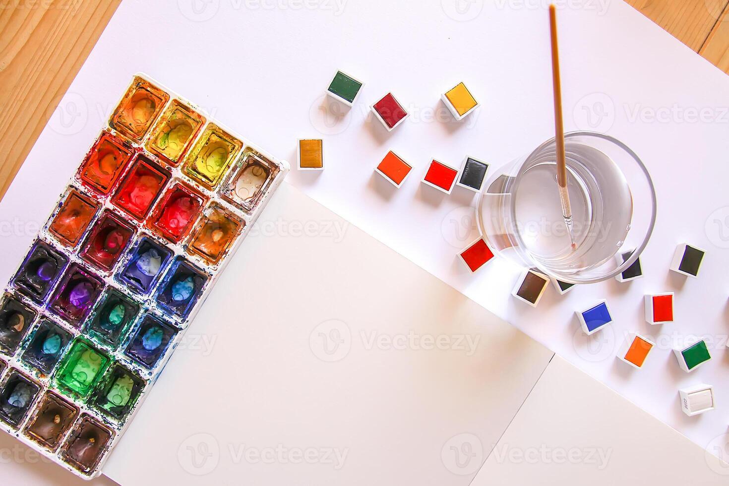 Watercolor paints, brushes, water in glass and paper on white background. Flat lay, top view photo