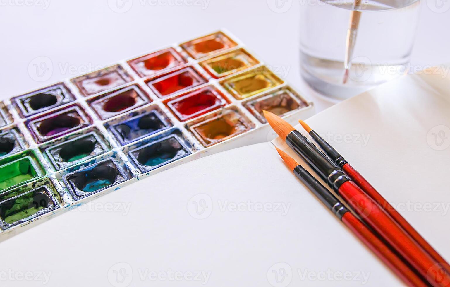 Watercolor paints, brushes, water in glass and paper on white background. Flat lay, top view photo