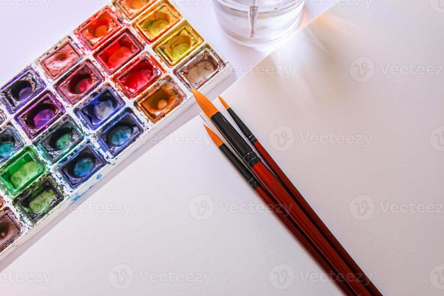 Watercolor paints, brushes, water in glass and paper on white background. Flat lay, top view photo