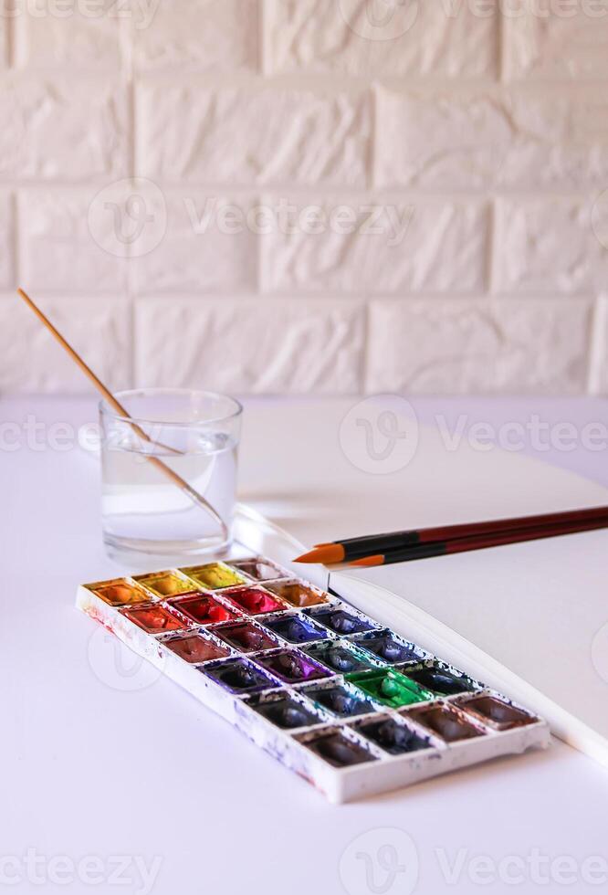 Watercolor paints, brushes, water in glass and paper on white background. Flat lay, top view photo