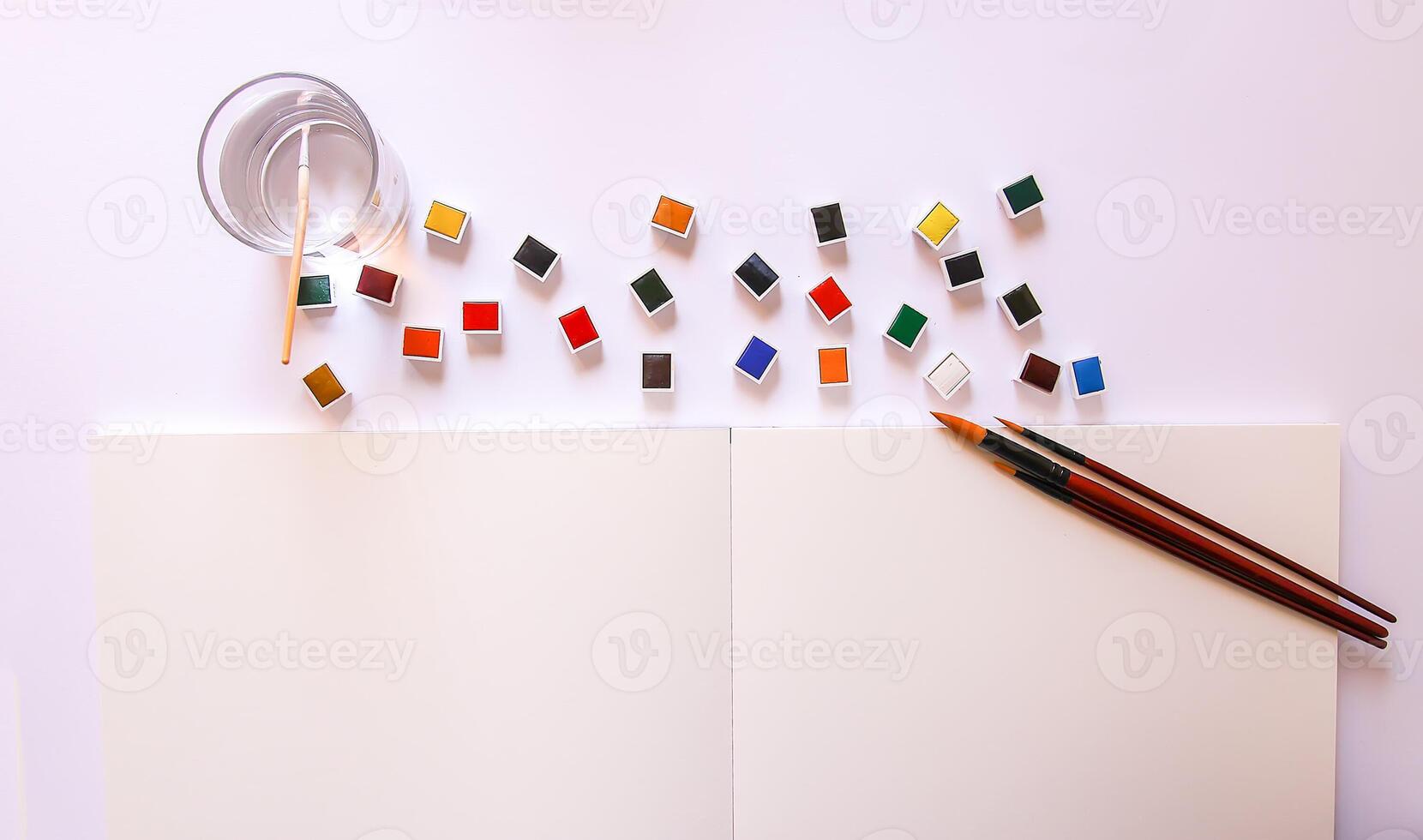 Watercolor paints, brushes, water in glass and paper on white background. Flat lay, top view photo