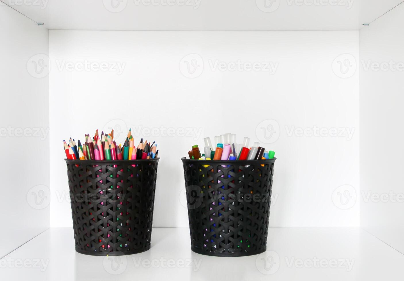 Colored crayons, pencils and pens in black containers on a white shelf. photo