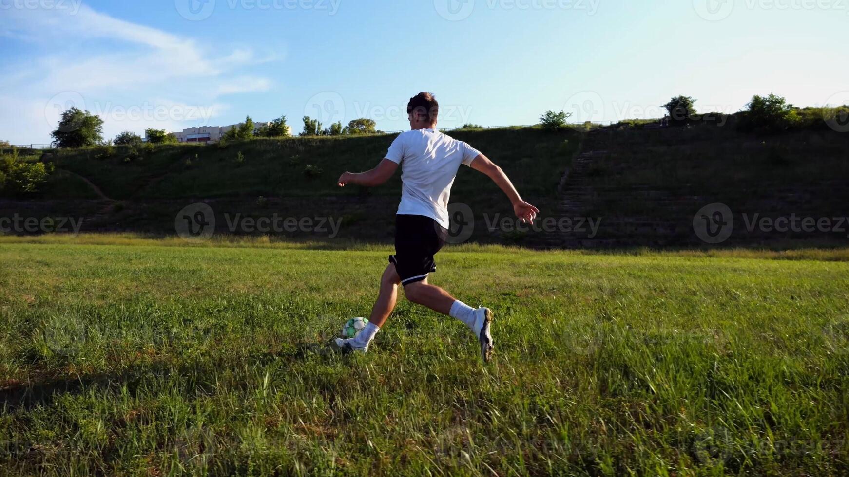 Professional footballer in uniform shows tricks with ball while running at green field. Sportsman kicks ball during jogging at meadow on sunny day. Young man training at stadium. Freestyle football photo