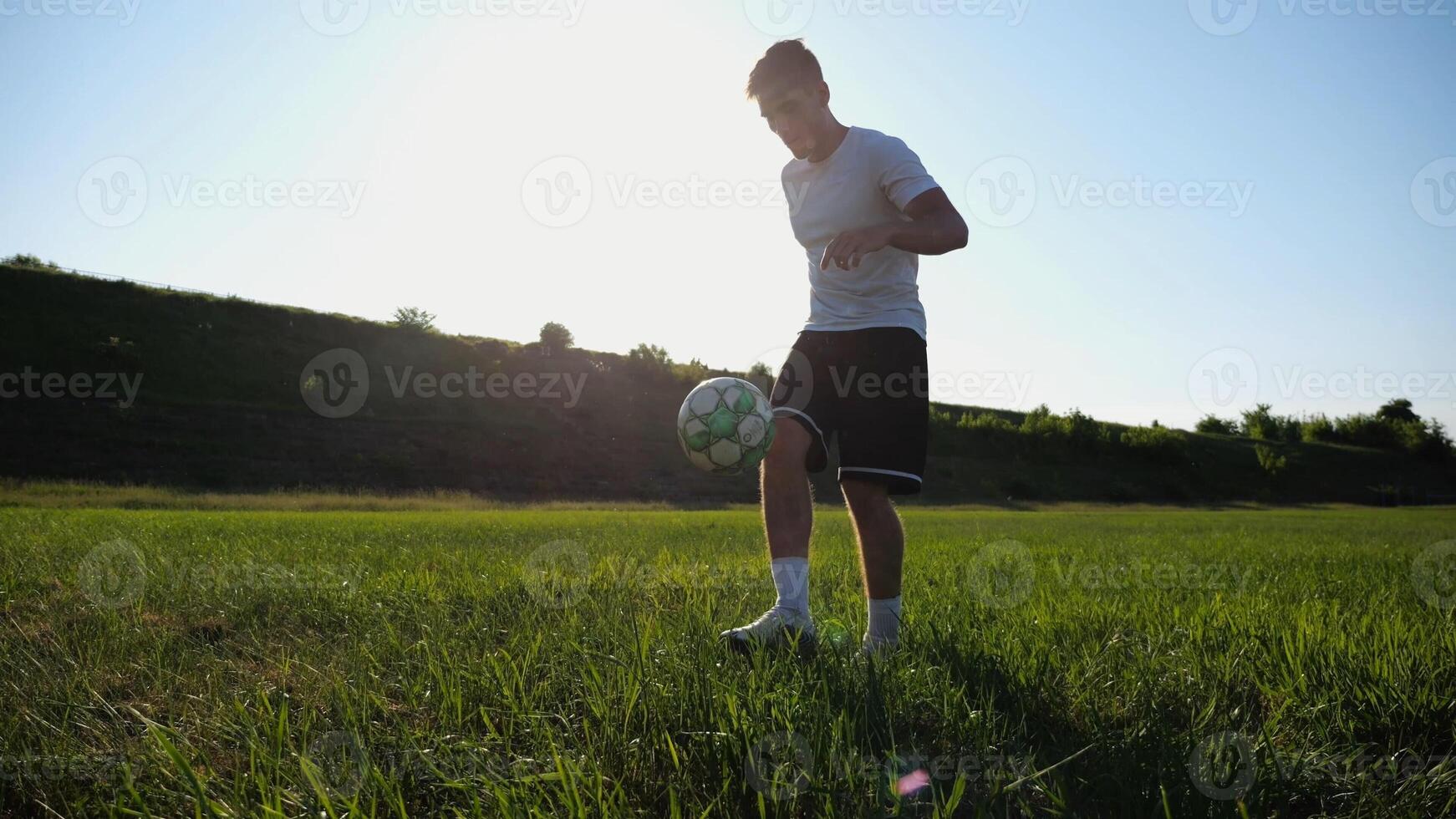 Sportsman juggling soccer ball on stadium at sunny day. Young man kicking ball at green field. Professional footballer practicing tricks at meadow with sunlight at background. Freestyle football. photo