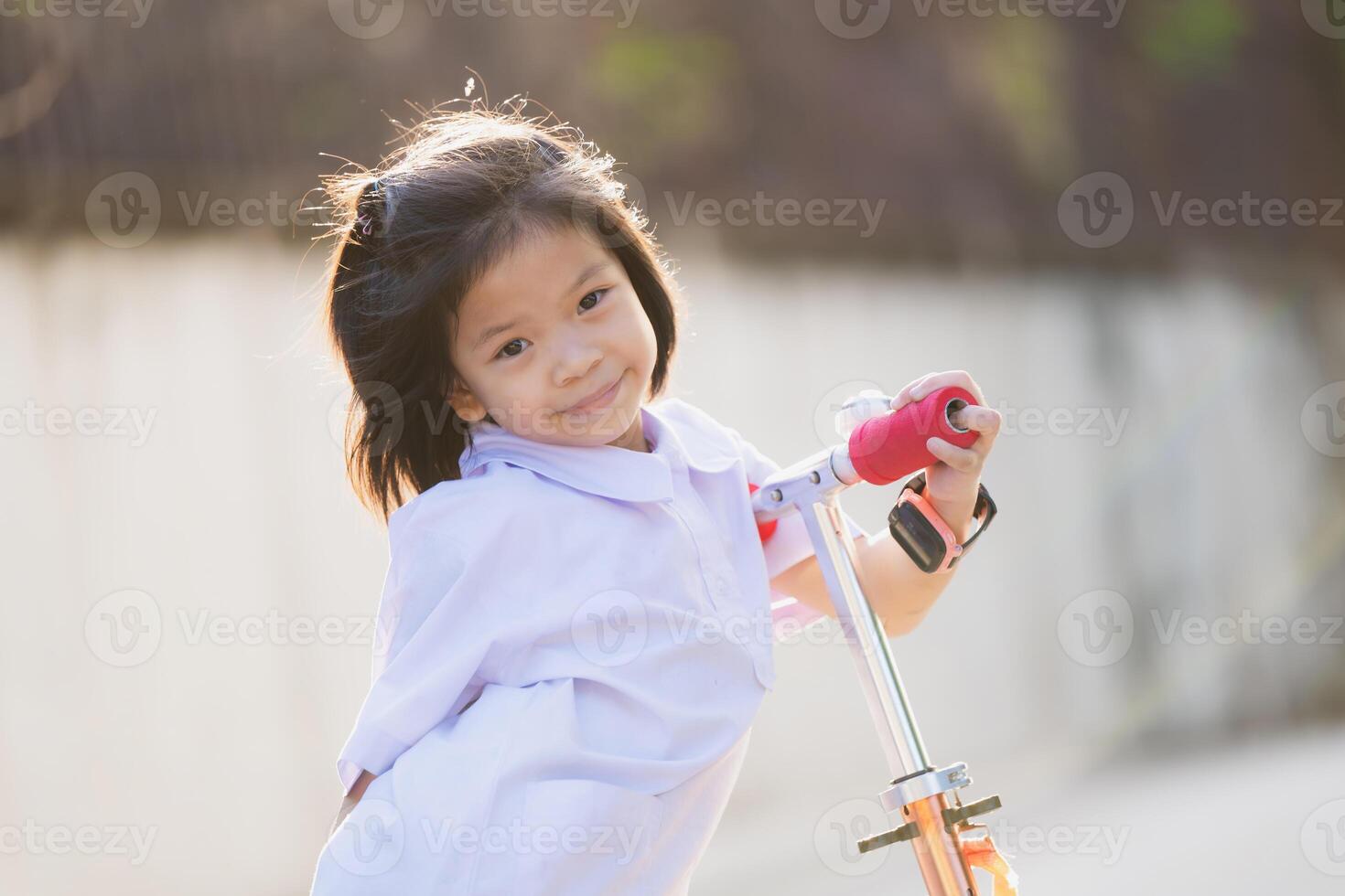Portrait of smiling child plays with red scootering outdoors in a park, sunny day. Girl playing on suburban street on summer or spring time. Healthy outdoor activity. Cute kid on his way to school. photo