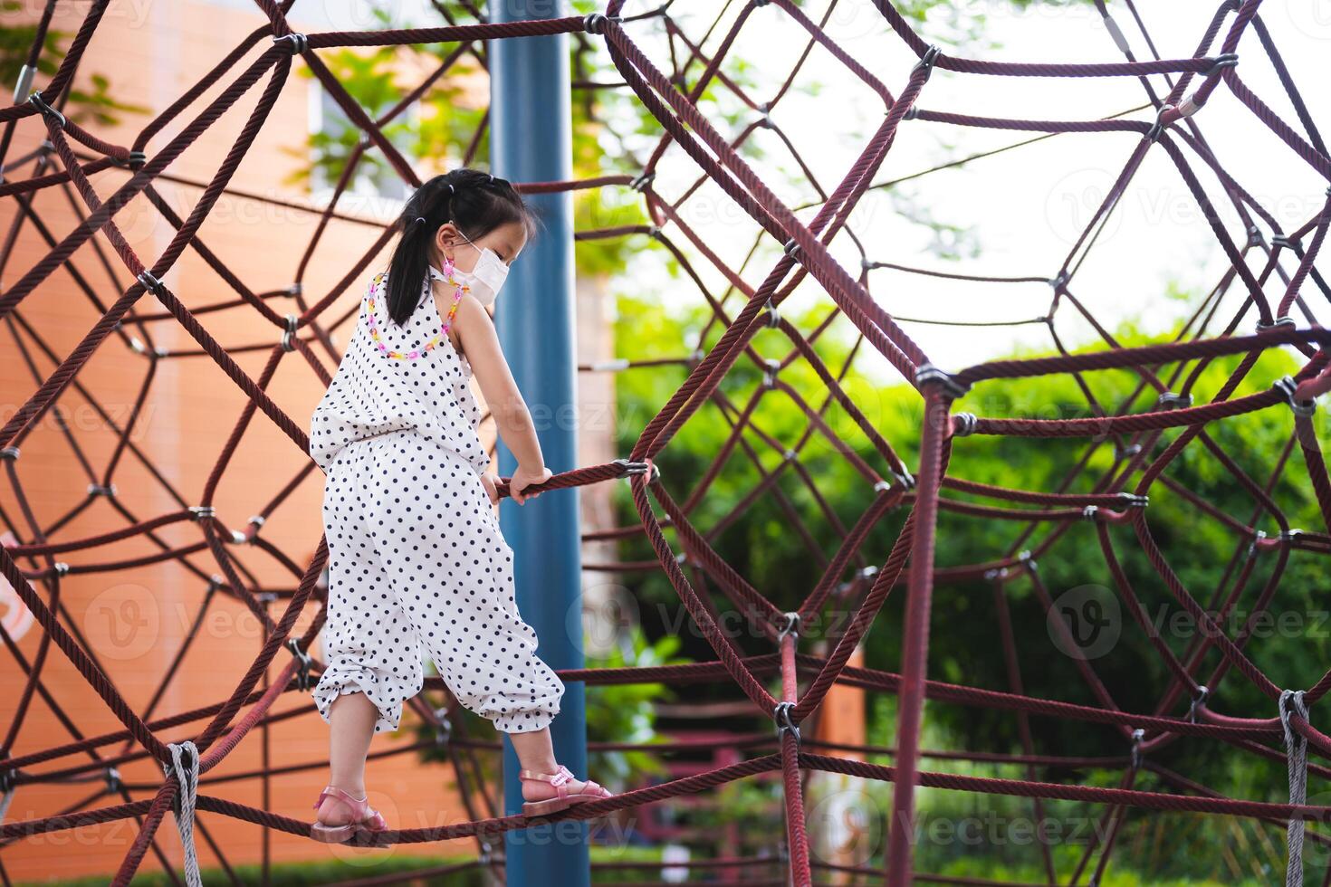 Girl having fun climbing a rope play equipment in playground. Child wear masks to protect her health. The play equipment is a rope that is woven into a three-dimensional shape for children to climb. photo