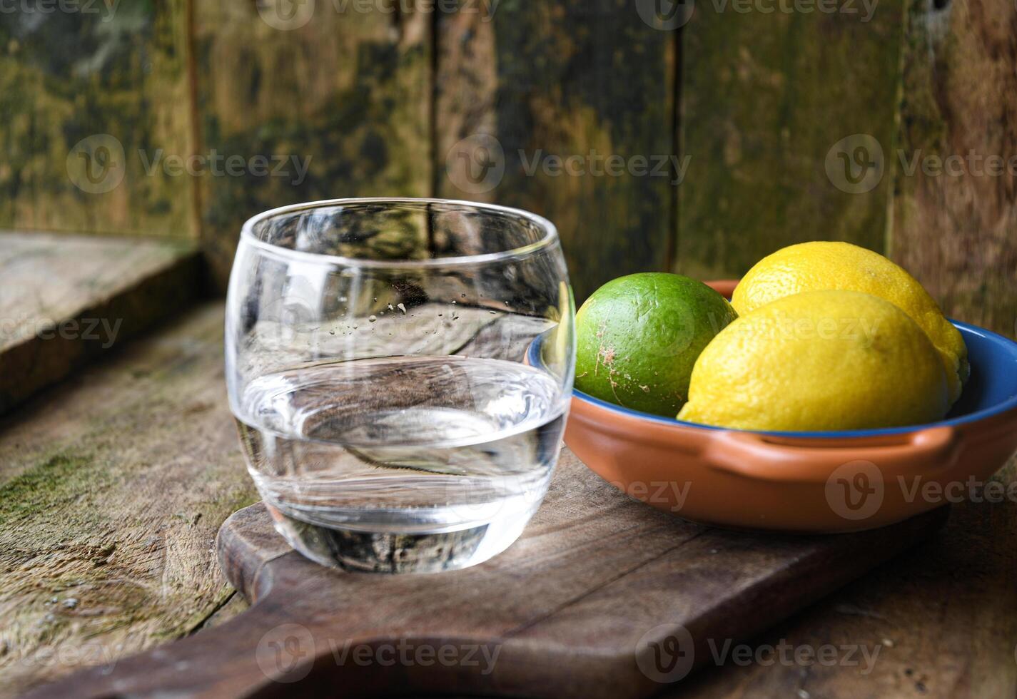 A glass of water and a ceramic plate with fruit, lemon, lime on a wooden table in a rustic kitchen. photo