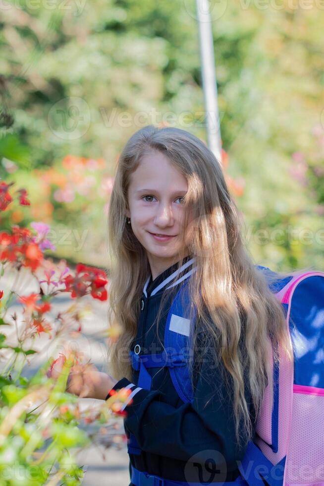 Back to school, portrait of a little girl from primary school outdoors, enjoying flowers in a flowerbed and looking at the camera with a smile, student dressed in dark blue uniform photo