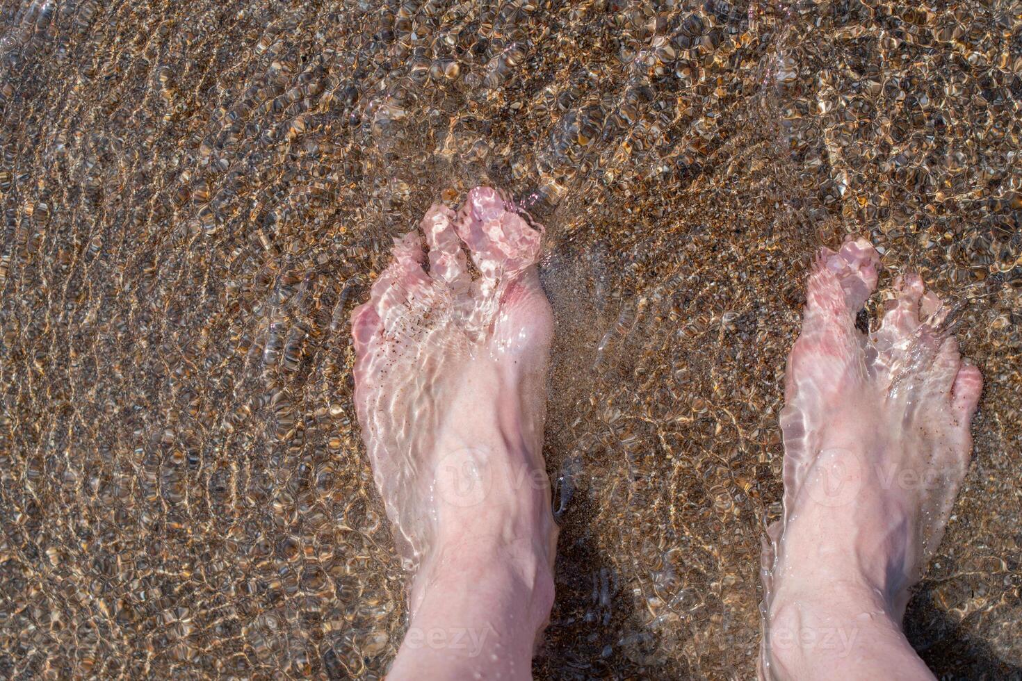 Female feet barefoot in waves of foamy water on a sandy beach on a summer day, top view of female feet, woman on the beach getting her feet wet in warm sea water, carefree vacation by the sea, photo
