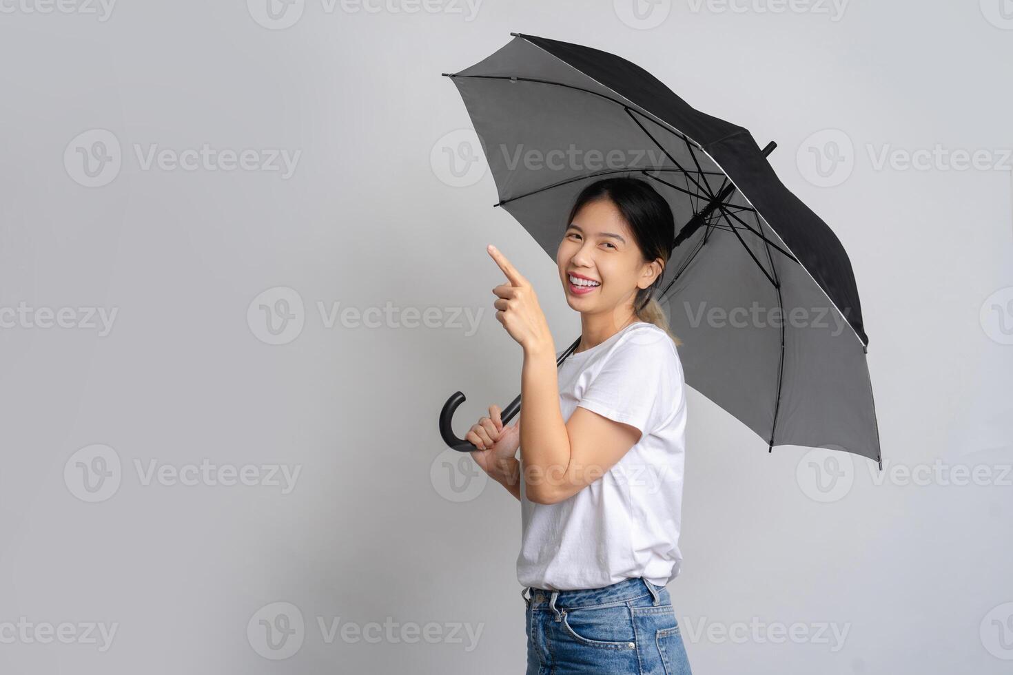 Happy Asian woman standing holding a umbrella and pointing at copy space on gray background. photo