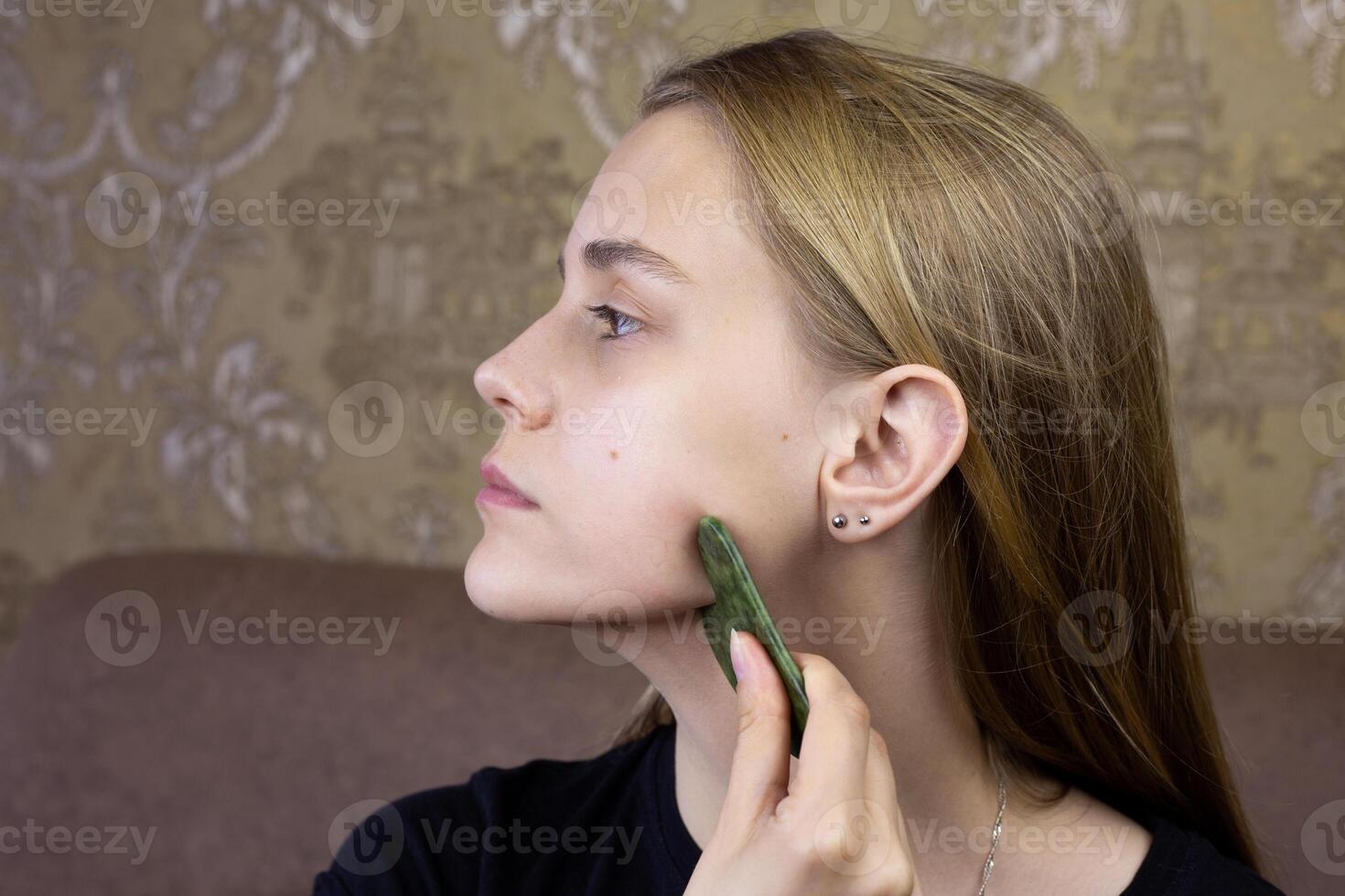 Profile of European girl who does facial massage using gua sha scraper to take care of health of skin and body, procedure for cleaning face and stretching wrinkles using special scraper photo
