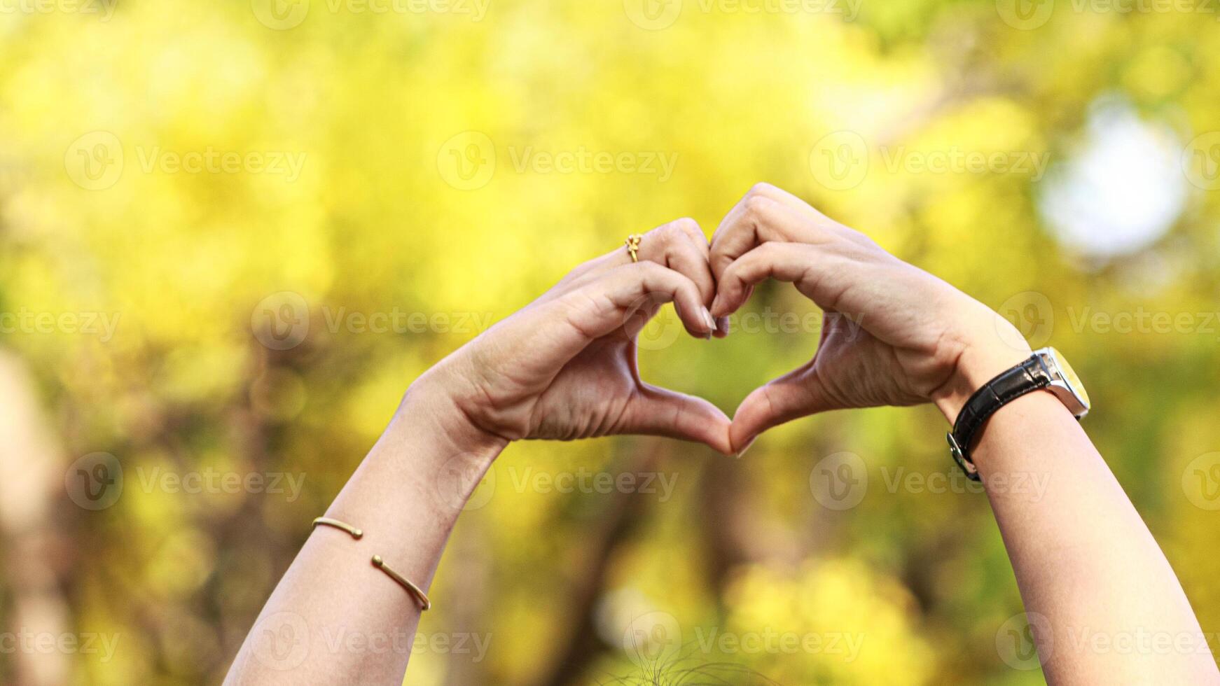 young woman raises her hands above to make heart shaped symbol that means friendship love and kindness on blurred background of beautiful morning nature Friendship and love concept photo