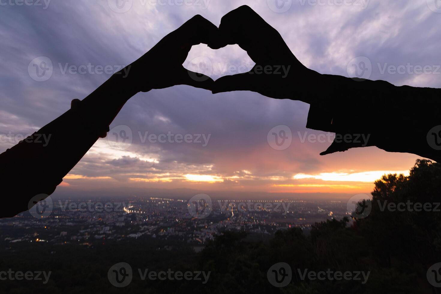 Silhouette of a pair of raised hands forming a heart shape on a beautiful evening landscape background Hand made heart symbol idea showing love friendship and kindness to couples and friends. photo