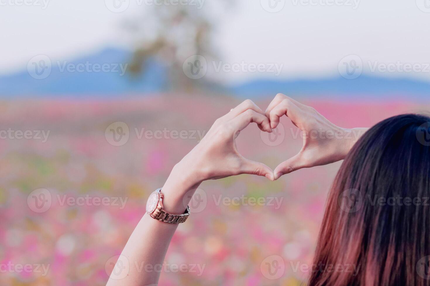 young woman raises her hands above to make heart shaped symbol that means friendship love and kindness on blurred background of beautiful morning nature Friendship and love concept photo