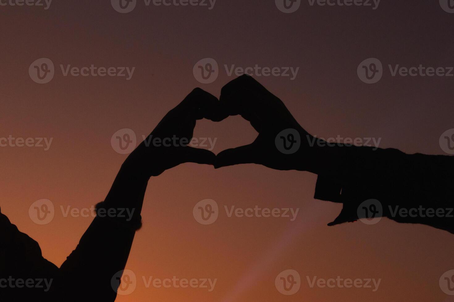 Silhouette of a pair of raised hands forming a heart shape on a beautiful evening landscape background Hand made heart symbol idea showing love friendship and kindness to couples and friends. photo
