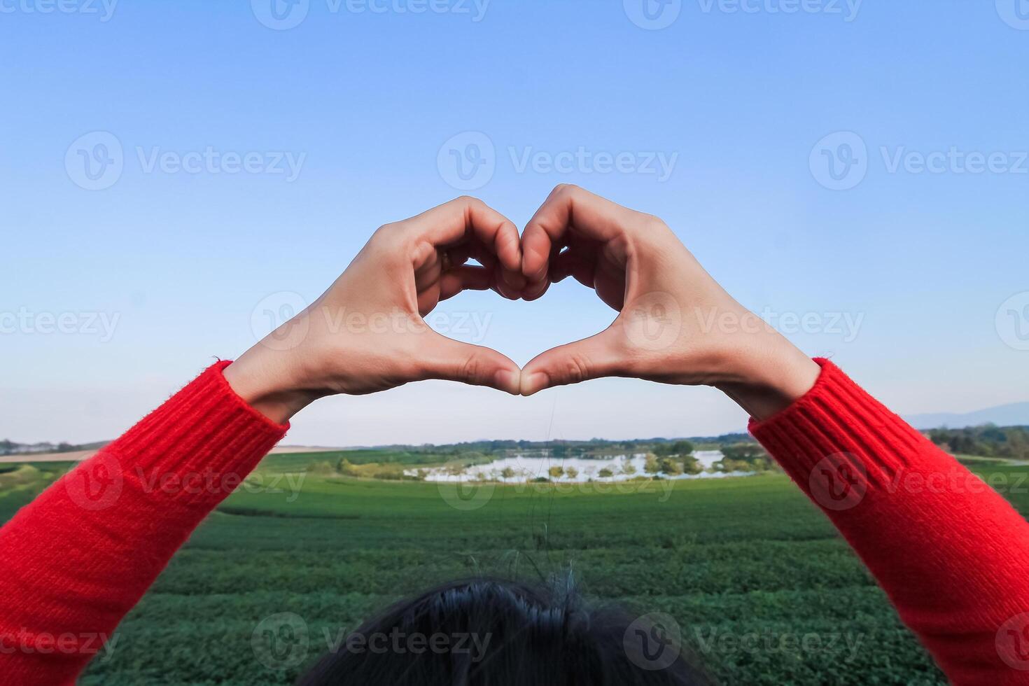 young woman raises her hands above to make heart shaped symbol that means friendship love and kindness on blurred background of beautiful morning nature Friendship and love concept photo