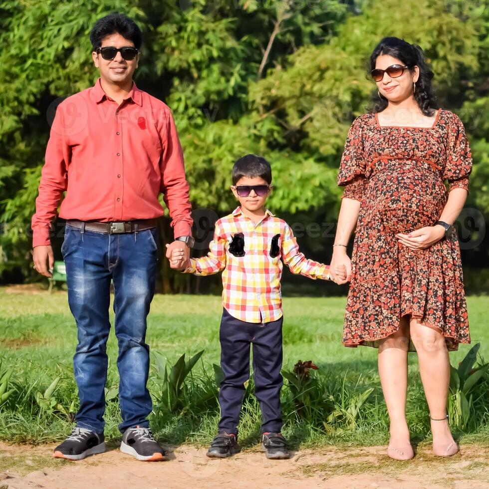 Indian couple posing for maternity baby shoot with their 5 year old kid. The couple is posing in a lawn with green grass and the woman is flaunting her baby bump in Lodhi Garden in New Delhi, India photo