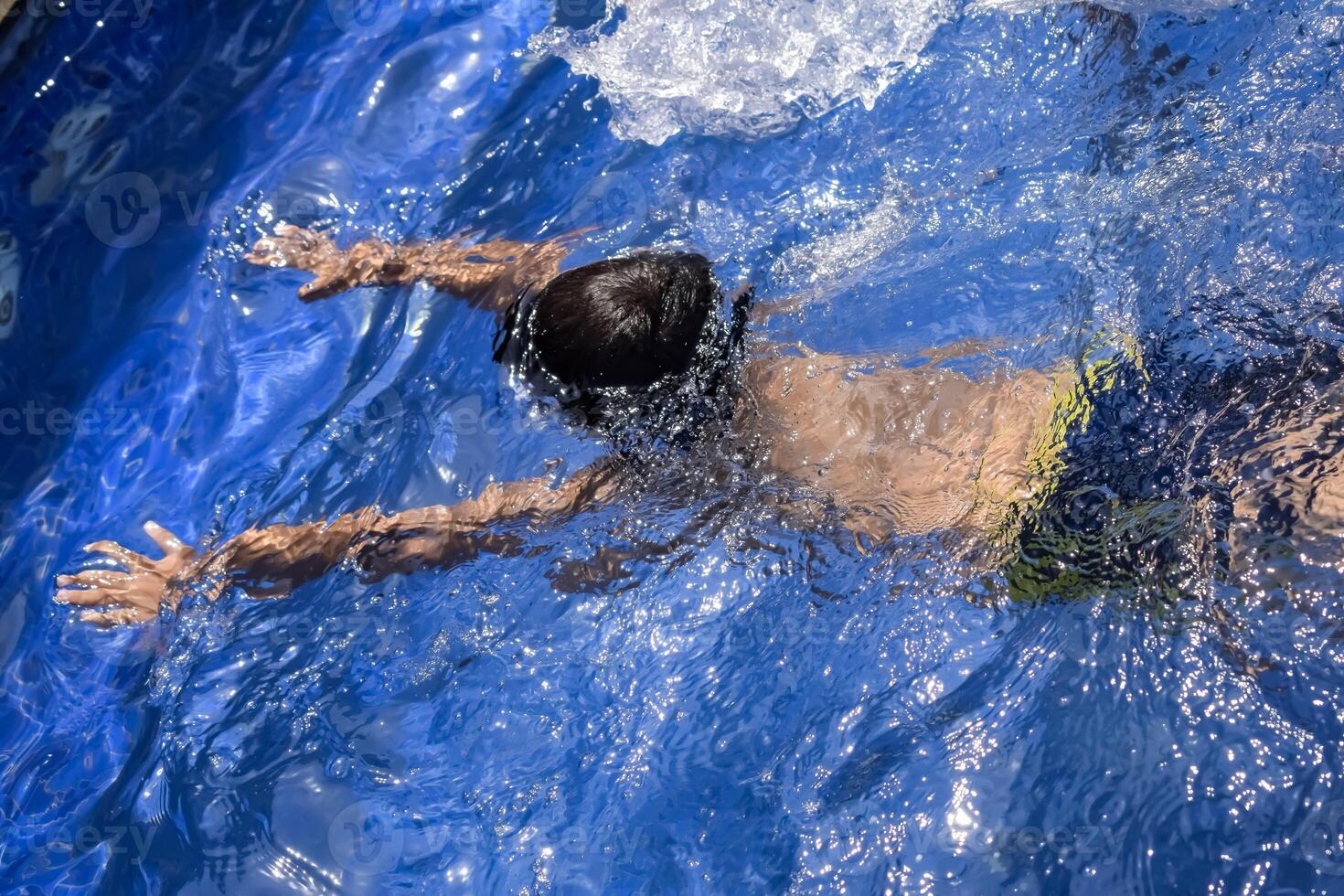 Happy Indian boy swimming in a pool, Kid wearing swimming costume along with air tube during hot summer vacations, Children boy in big swimming pool. photo