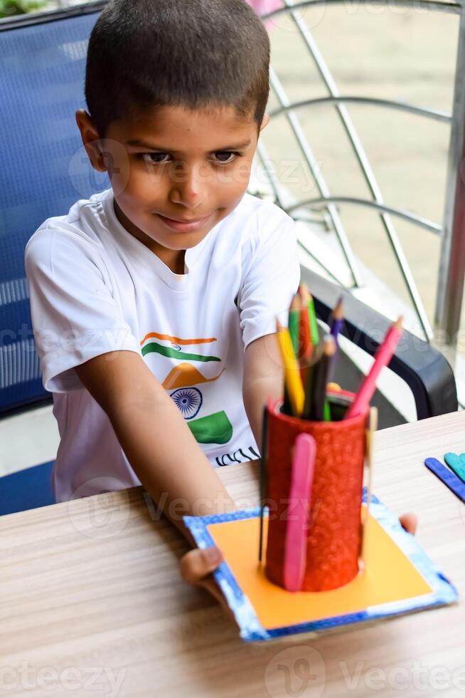 Smart Indian little boy performing best out of waste activity with different colourful objects during the summer vacations, Cute Indian Kid doing best our of waste activity at home on wooden table photo